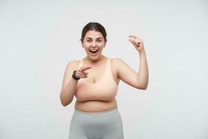 Portrait of young joyful dark haired oversized female demonstrating small size with raised fingers and looking cheerfully at camera with wide smile, isolated over white background photo