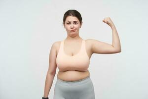 Indoor shot of young confused chubby lady with ponytail hairstyle twisting her mouth while looking at camera with pout, isolated over white background. Concept of determination will-power photo