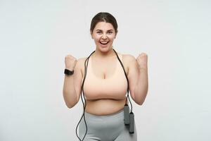 Indoor shot of cheerful young dark haired oversized woman with skipping rope raising happily her fist while rejoicing about nice working out, isolated over white background photo