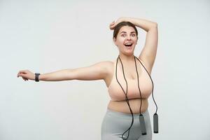 Indoor photo of joyful young dark haired chubby lady with ponytail hairstyle stretching her hands and looking excitedly at camera, posing over white background