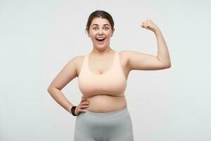 Overjoyed young lovely dark haired oversized female dressed in sporty wear looking happily at camera while demonstrating her power over white background. Concept of weight loss photo