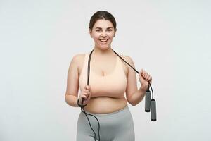 Cheerful young lovely plus size model with ponytail hairstyle wearing jumping-rope on her neck and smiling gladly at camera, being pleased after hard training. Body positive concept photo
