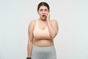 Frightened young oversized woman with ponytail hairstyle looking scaredly at camera with wide mouth opened and raising hand to her face, standing over white background photo