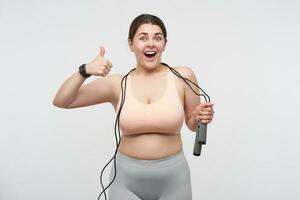 Agitated young dark haired plump woman dressed in sporty clothes wearing jumping-rope and showing happily thumb to camera while posing over white background. Concept of weight loss photo