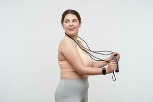 Positive young lovely dark haired chubby lady dressed in sports bra and leggins wearing jumping-rope on her neck and looking cheerfully over her shoulder. Body positive concept photo