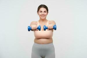 Studio shot of young pleased oversized female making static exercises with weight and smiling gladly at camera, working out her body while posing over white background photo