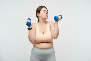 Positive young oversized female dressed in sports bra and leggins keeping blue dumbbells in raised hands and forming lips in kiss while looking at it, isolated over white background photo