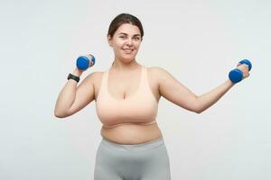 Cheerful young cute oversized woman in sporty clothes smiling nicely at camera while training her arms with blue dumbbells, posing over white background. Concept of weight loss photo