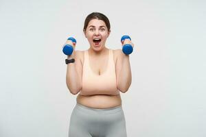 Indoor photo of young brunette plump lady with chubby face lifting dumbbells and looking excitedly at camera while posing over white background. Bodycare concept