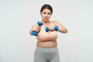 Studio shot of young plump oversized woman with ponytail hairstyle keeping dumbbells in raised hands while making fitness at home, isolated over white background photo