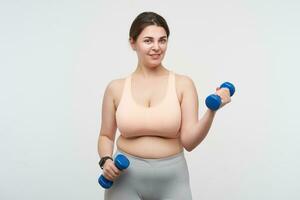 Cute young brunette chubby plump lady dressed in sporty clothes working out her hands with dumbbells and looking positively at camera while standing over white background photo