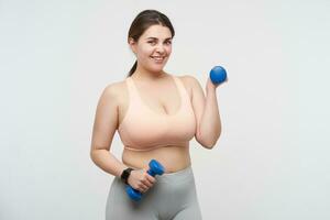 Positive young plus size model with ponytail hairstyle making sports while posing over white background and smiling gladly at camera. Body positive concept photo