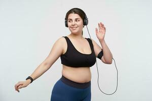 Glad of young brown haired chubby lady dressed in sports bra and leggins smiling positively while working out to music in her earphones, isolated over white background photo