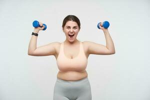 Excited young brown haired chubby female dressed in sporty top and leggins raising emotionally hands with dumbbells while posing over white background. Concept of self-made determination will-power photo