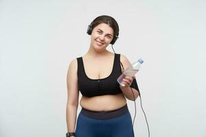 Cheerful young brunette fatty female with chubby face smiling happily at camera while preparing for working out and listening to music in headphones, standing over white background photo