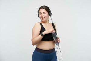 Positive young lovely brunette oversized female drinking water while having fitness class and being in high spirit, posing over white background. Body positive concept photo