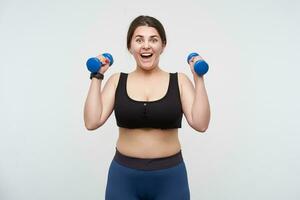 Agitated young cheerful oversized woman with casual hairstyle looking emotionally at camera while raising hands with blue dumbbells, posing over white background. Body positive concept photo