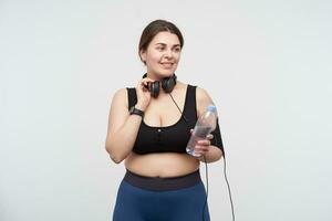 Glad young lovely brunette oversized lady with casual hairstyle drinking water after morning workout and smiling cheerfully, wearing earphones on her neck while posing over white background photo