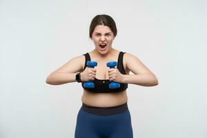 Expressive young brunette oversized lady dressed in sports bra and leggins looking emotionally at camera during hard training, isolated over white background. Concept of weight loss photo