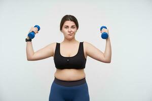 Puzzled young oversized woman with casual hairstyle looking confusedly at camera and twisting mouth while making exercises with dumbbells, posing over white background photo