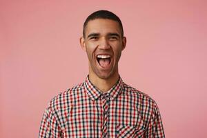 Portrait of laughing attractive dark skinned guy in checkered shirt, looks at the camera with happy expression and wide open mouth, hears funny joke, stands over pink background. photo