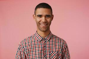 Close up of young happy attractive dark skinned guy in checkered shirt, looks at the camera with positive expression, broadly smiling and stands over pink background. photo