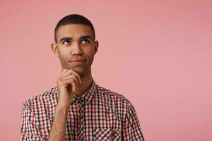 young pensive attractive dark skinned guy in checkered shirt, looks away and touches chin, thinks about the task list for tomorrow, stands over pink background with copy space at the right side. photo