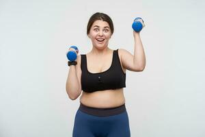 Overjoyed young brunette plump female dressed in sporty wear training her arms using little blue dumbbells while looking excitedly at camera, posing over white background photo