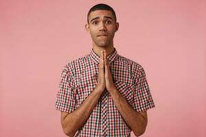 Portrait of young attractive dark skinned guy in checkered shirt, cupped hands together in pray gesture, bites lip, look at the camera and hopes for luck,stands over pink background. photo