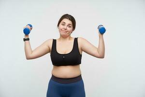 Indoor shot of confused young chubby female dressed in sporty clothes training her arms with blue dumbbells while standing over white background. Concept of determination will-power photo