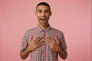 Portrait of happy amazed young attractive dark skinned guy in checkered shirt, looks at the camera with wide open mouth and eyes, stands over pink background with palms on on rude. photo