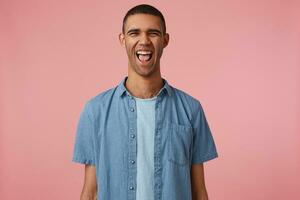 Laughing young attractive dark skinned guy in checkered shirt, looks at the camera with happy expression and wide open mouth, hears funny joke, stands over pink background. photo