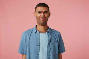 Portrait of frowning disgusted dark skinned boy with clenched lips, saw something repulsive and unpleasant, stands over pink background. People and emotion concept. photo