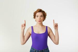 Portrait of a young happy amazed attractive short-haired girl, wearing a purple jersey, looking at the camera, speaks lips wow and index fingers point up at the copy space isolated over white wall. photo