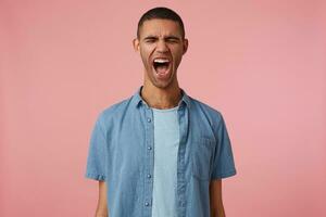 Portrait of young screaming angry dark skinned boy with closed eyes, stands wit wide open mouth over pink background. People and emotion concept. photo