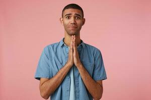 Young attractive dark skinned guy in checkered shirt, cupped hands together in pray gesture, look at the camera and hopes for luck,stands over pink background. photo