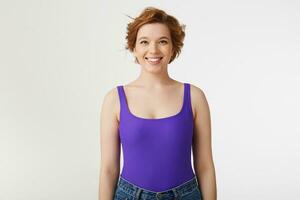 Portrait of a young attractive short-haired girl, wearing a purple jersey and jeans, smiling broadly and looking at the camera isolated over white wall. photo