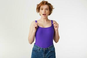 Young surprised attractive short-haired girl, wearing a purple jersey, looking at the camera with open eyes and mouth isolated over white wall. photo