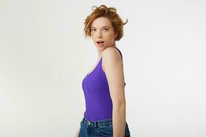 Young surprised attractive short-haired girl, wearing a purple jersey,posing in profile, looking at the camera with wide open mouth and eyes, she heard the news. Isolated over white wall. photo