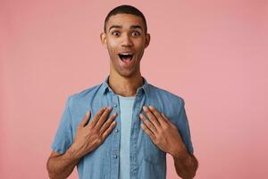 Happy amazed young attractive dark skinned guy in blank shirt, looks at the camera with wide open mouth and eyes, stands over pink background with palms on on rude. photo