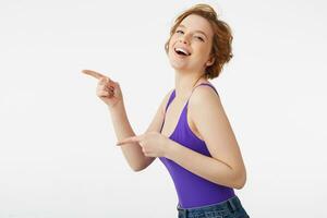 Portrait of a young attractive smiling short-haired girl, wearing a purple jersey and jeans, winking and looking at the camera, laughs, points the finger at copyspace isolated over white wall. photo