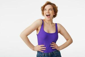 Photo of young happy surprised attractive short-haired girl, wearing a purple jersey, looking up with open eyes and mouth isolated over white wall with copy space.