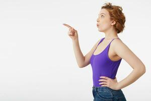 Happy amazed young attractive short-haired girl, wearing a purple jersey, saw something incredibly interesting and points the finger, look at copy space isolated over white wall. photo