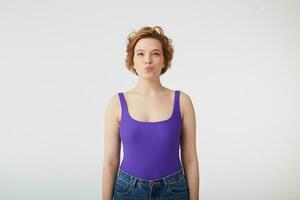 Portrait of young cute short-haired girl, wears in purple shirt and jeans, looks at the camera and keeps lips folded, stands over white background. photo