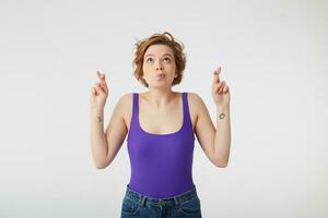 Portrait of happy young attractive short-haired girl, fingers crossed and lips pressed upwards and looks forward to good luck in the chemistry exam, stands over white wall. photo