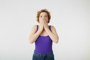 Photo of amazed young attractive curly short-haired girl, heard the incredible news and covers her mouth with hands in surprise, stands over white wall.