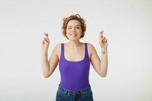 Portrait of happy young attractive short-haired girl, winks and bites lip, fingers crossed hoping for the fulfillment of the wish, stands over white wall. photo