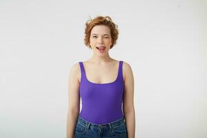 Photo of happy funny young cute short-haired girl, winks and shows tongue, wears in purple shirt, stands over white background.