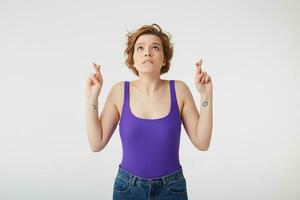 Photo of young nice short-haired girl, fingers crossed and lips pressed upwards and looks forward to good luck in the exam, stands over white wall.