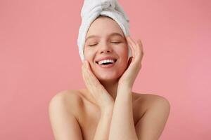 retrato de joven contento mujer después ducha con un toalla en su cabeza, en general sonrisas con cerrado ojos, toques cara y suave piel, soportes terminado rosado antecedentes. foto